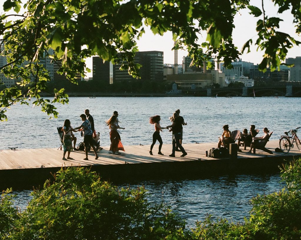 Framed by green leaves, a group of people dance together on an Esplanade pier with the Cambridge skyline in the background.