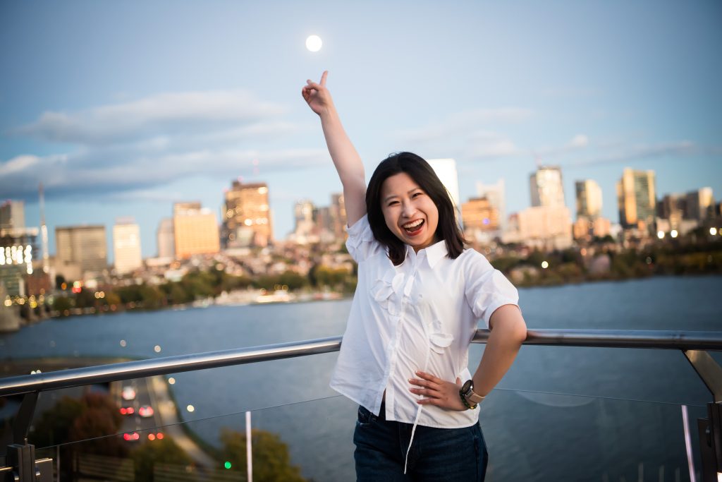 A Presidential Fellow smiles wide, pointing at the moon above the Boston skyline.