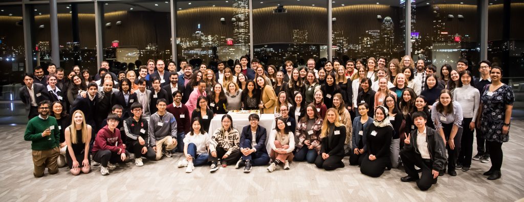 A group photo of all of the Presidential Fellows with Chancellor Nobles, a 25th Anniversary cake in the center of the photo and the Boston skyline at night in the background.