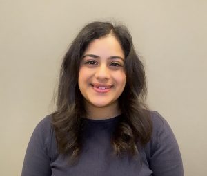 a dark haired graduate student smiles against a plain taupe background.